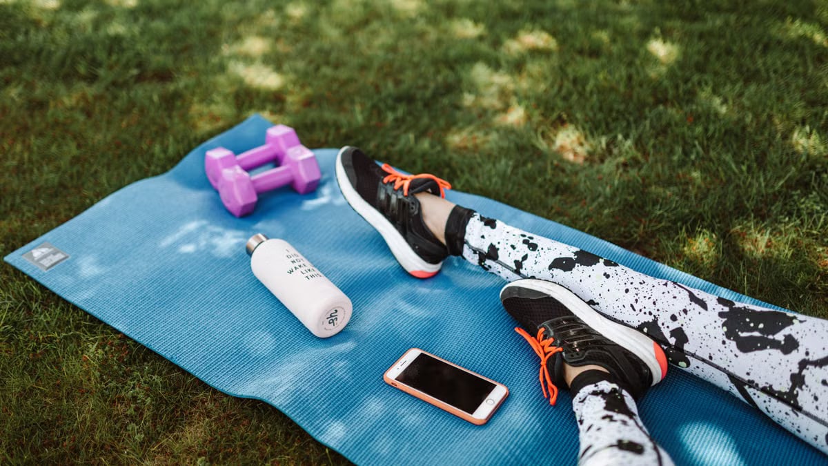 Focused workout setup outdoors with mat, dumbbells, phone, and water bottle.